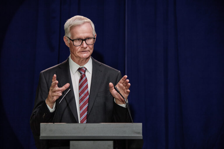 Medium shot of senior politician in formal attire giving public speech announcing legislative initiative during press conference while standing at podium on stage against blue curtains, copy space