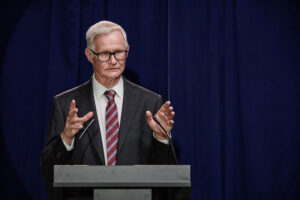 Medium shot of senior politician in formal attire giving public speech announcing legislative initiative during press conference while standing at podium on stage against blue curtains, copy space